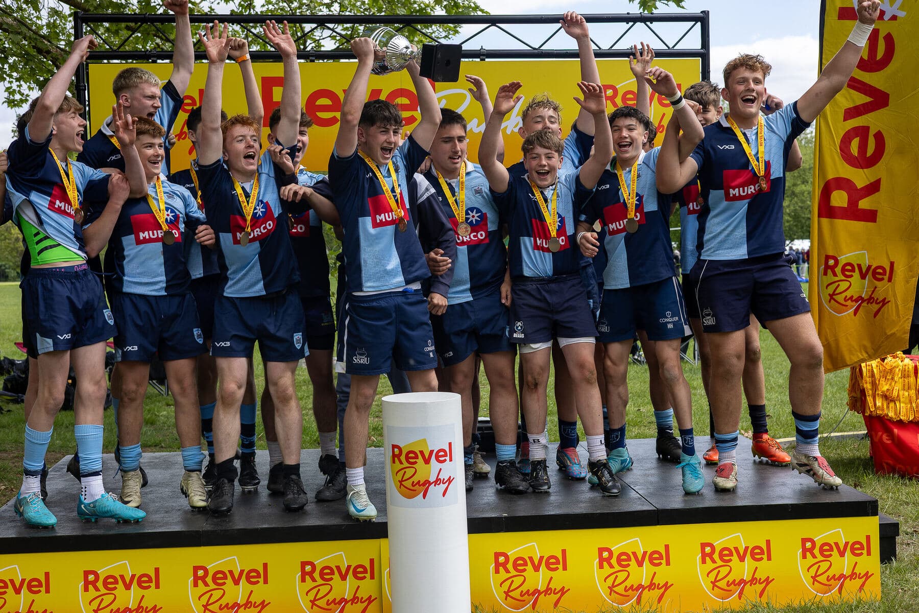 A junior squad in navy and sky blue celebrating on the Revel podium, medals on and the cup held high