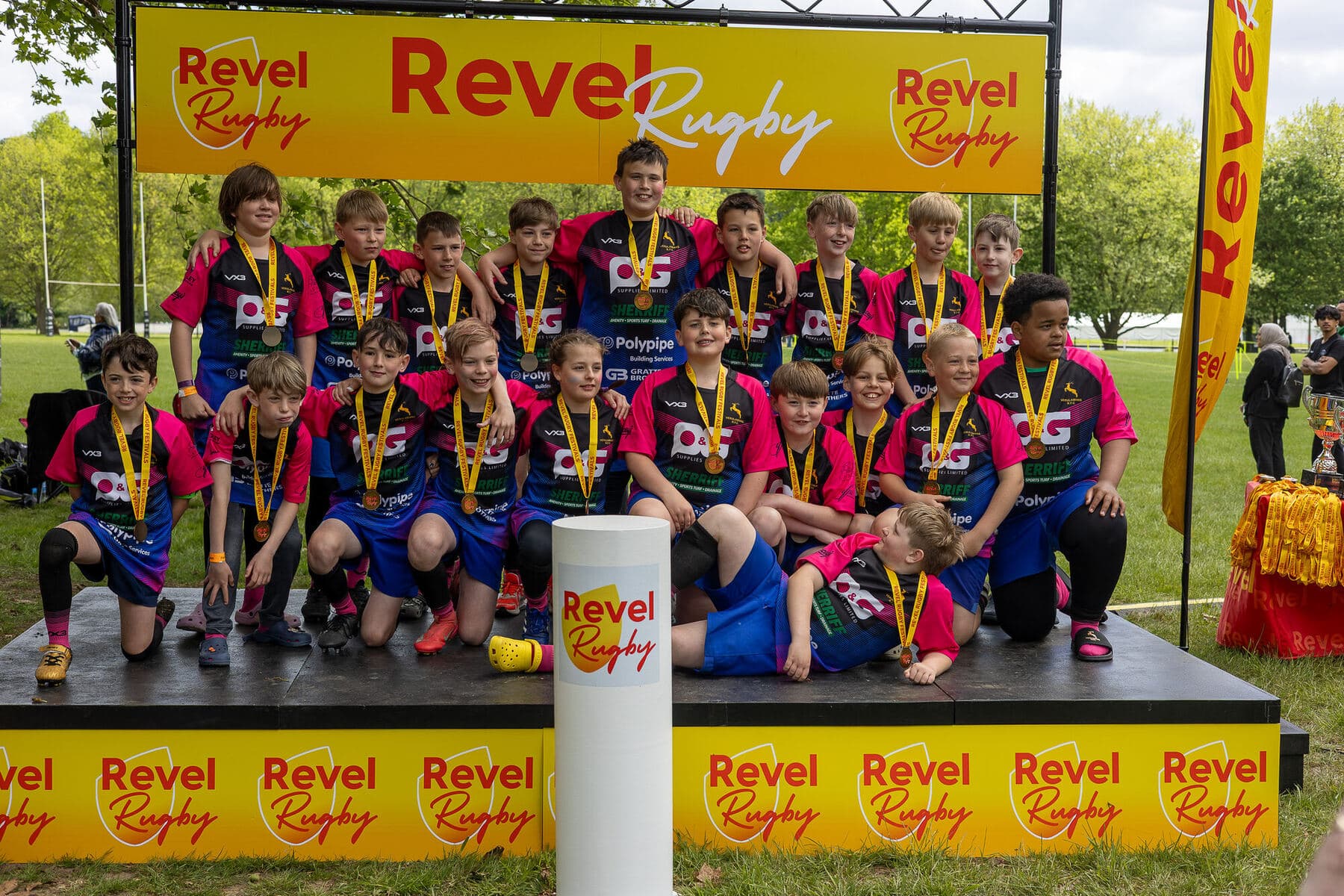 A junior squad in pink and blue kit gathered on the Revel Rugby podium with medals round their necks, the trophy table beside them