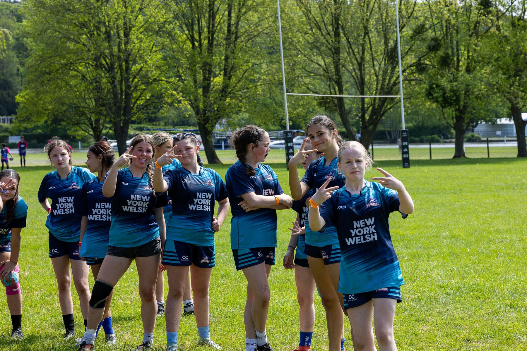 A girls team in teal New York Welsh shirts standing together on a sunlit pitch, laughing and posing for the camera