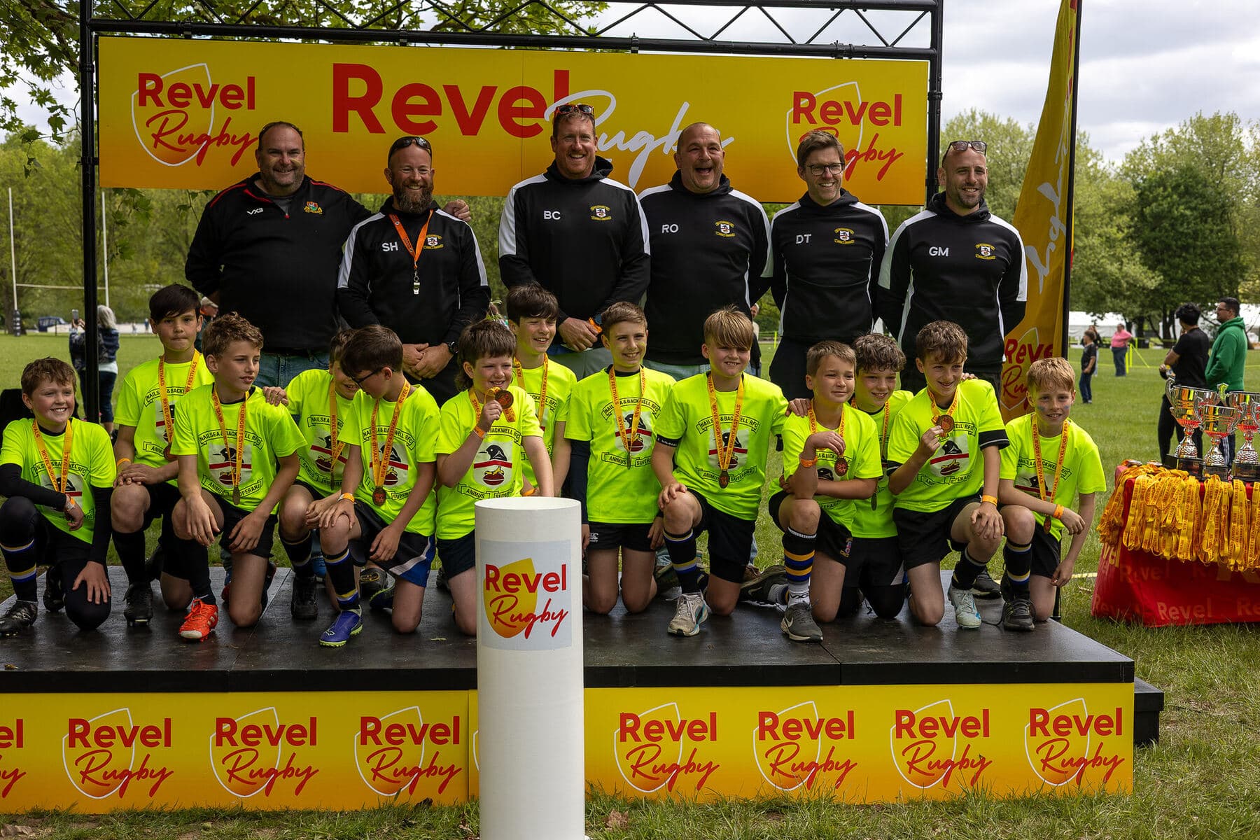 A squad in bright yellow shirts kneeling with medals round their necks on the Revel Rugby podium, six coaches and staff standing behind them