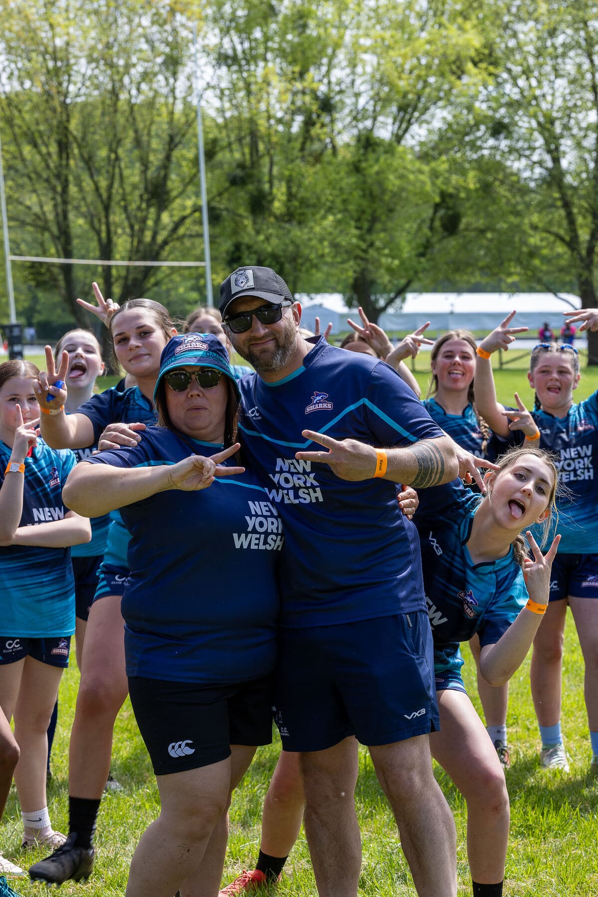 A girls squad crowding in behind their two coaches on the pitch, everyone mugging for the camera in the sun