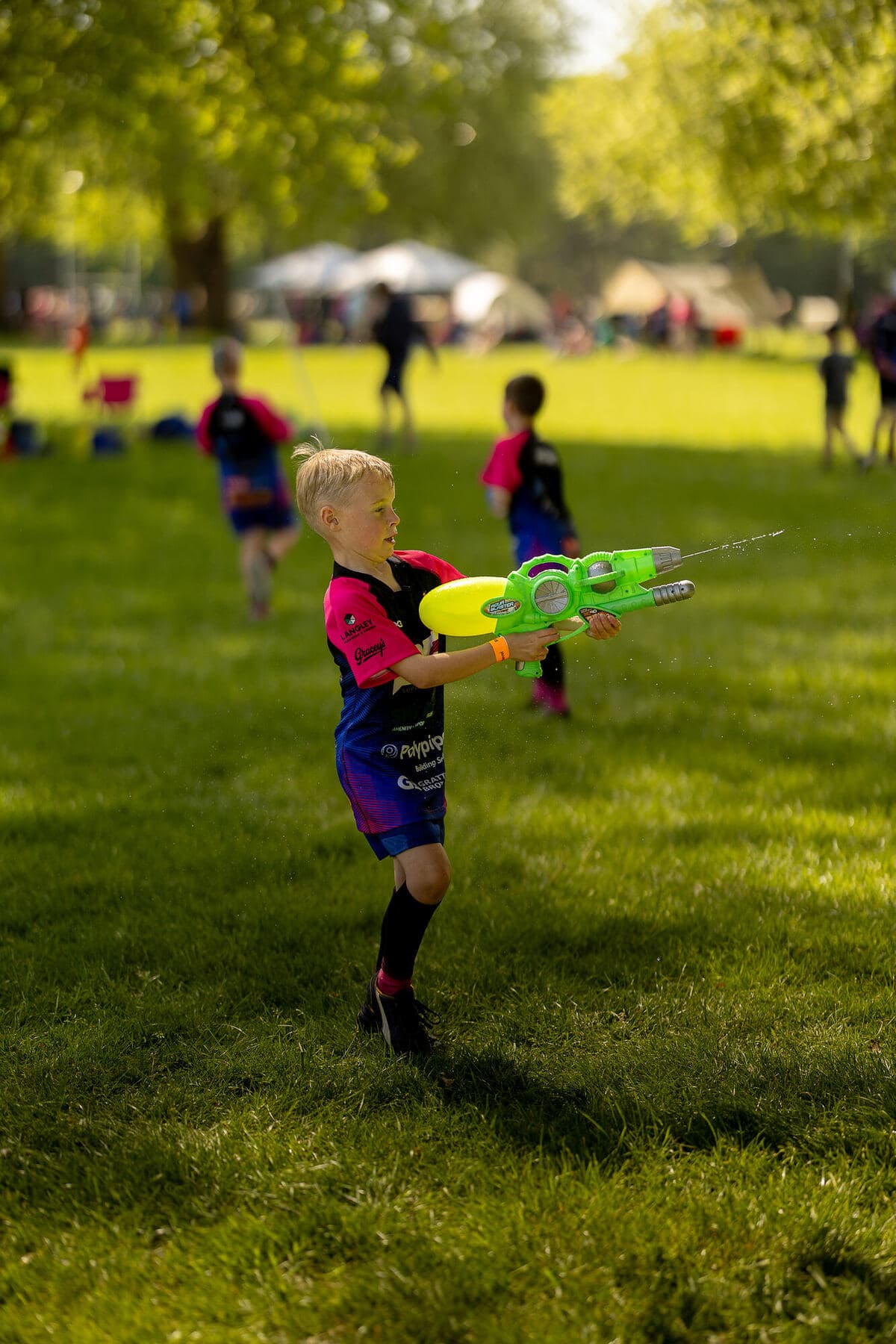 A young player in club kit playing with a water pistol in the sunshine on the grass between matches, families and gazebos behind him