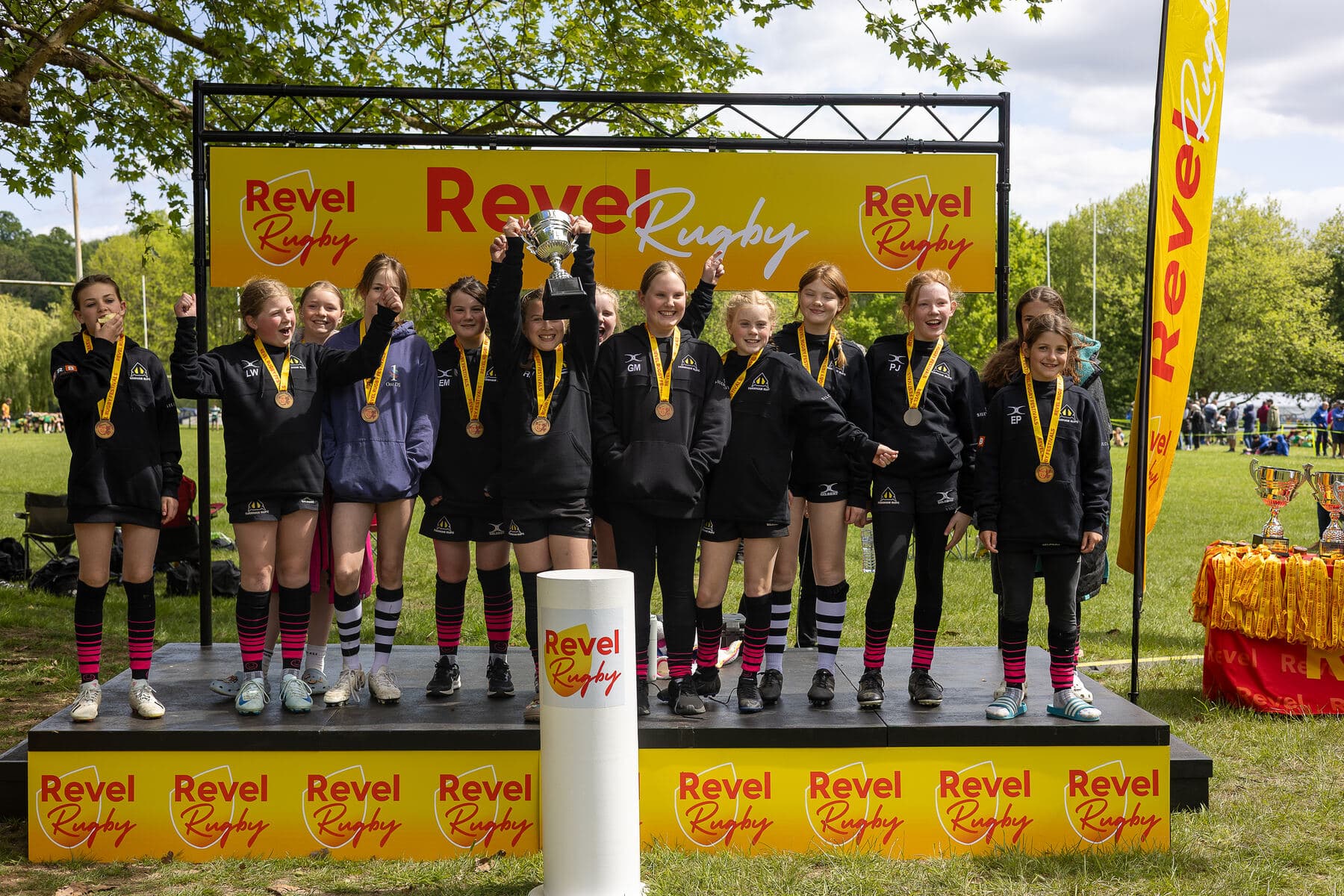 A girls team lined up on the Revel Rugby podium with medals on, one player holding the cup above her head