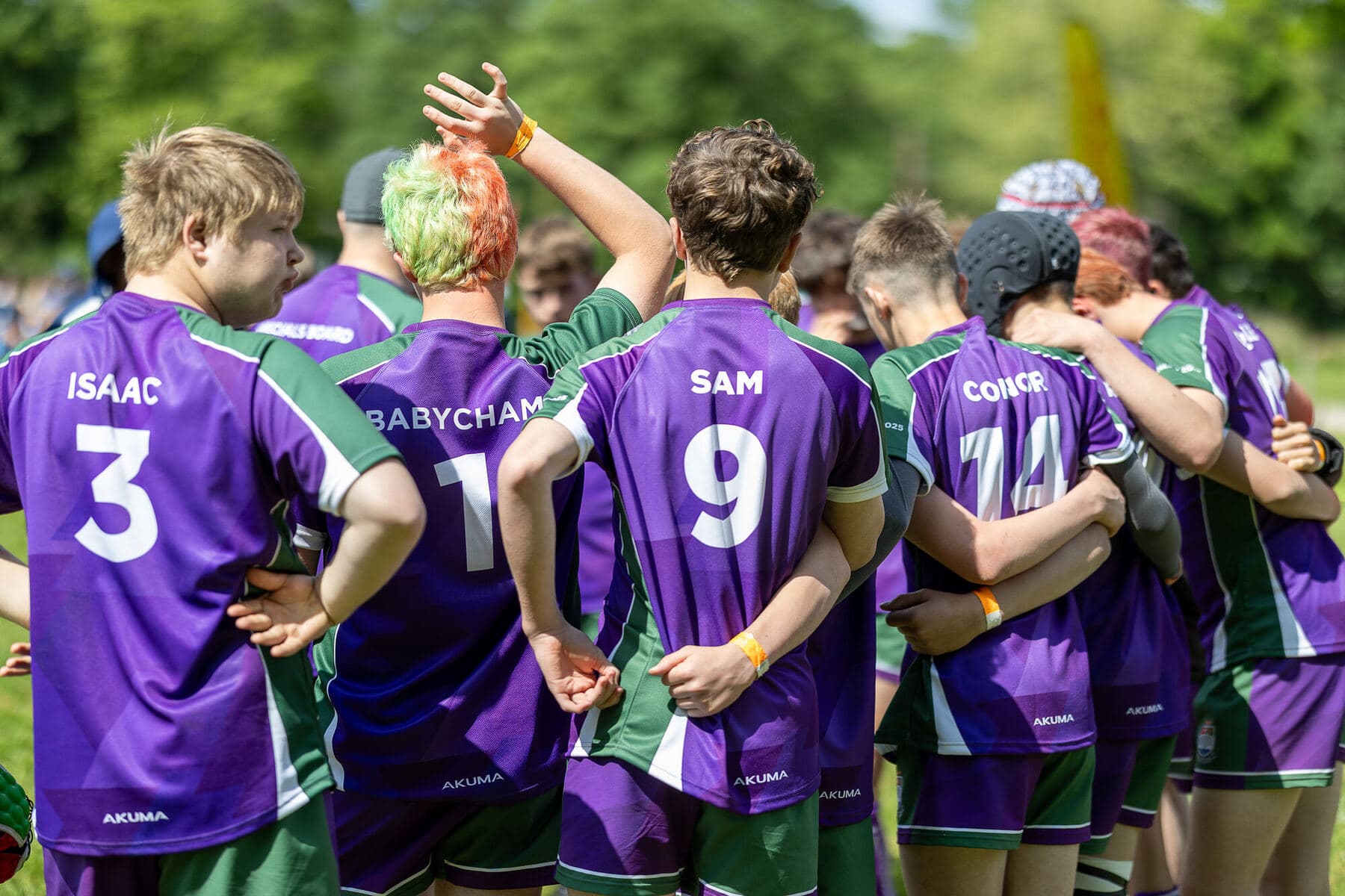 An Under 16 squad in a huddle in the sunshine, first names printed across the backs of their purple and green shirts