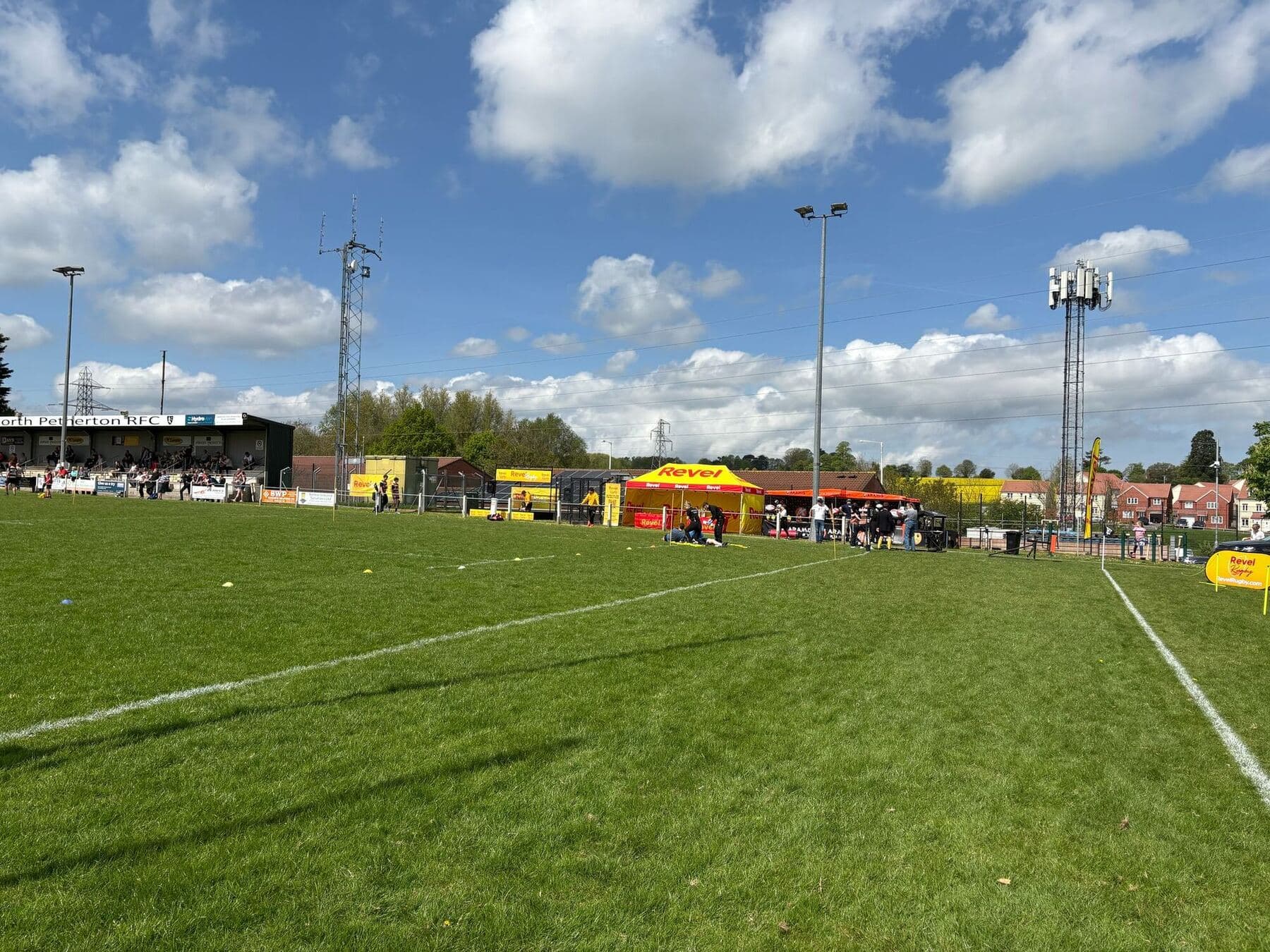 A full-size pitch marked out in front of a clubhouse stand under a bright sky
