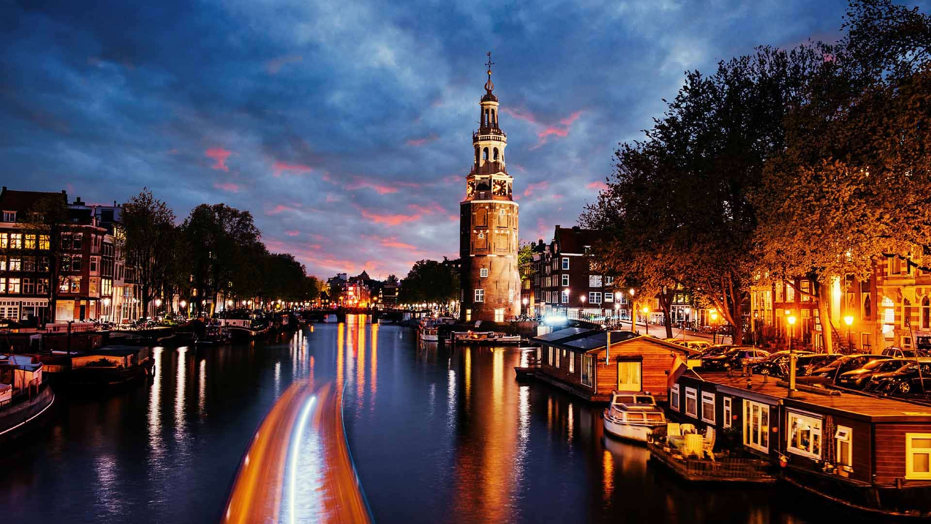 An old clock tower above an Amsterdam canal at dusk, houseboats lit up along the water