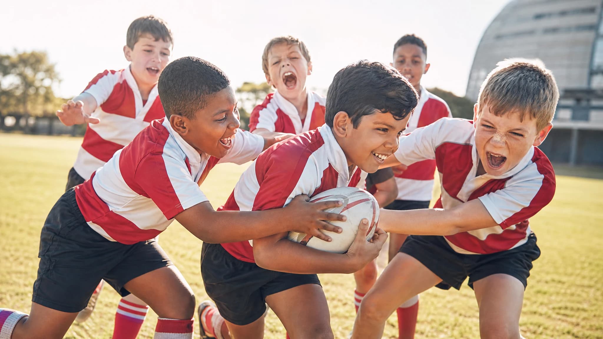Minis rugby players in red and white hoops laughing as they carry the ball into a tackle on a sunny pitch
