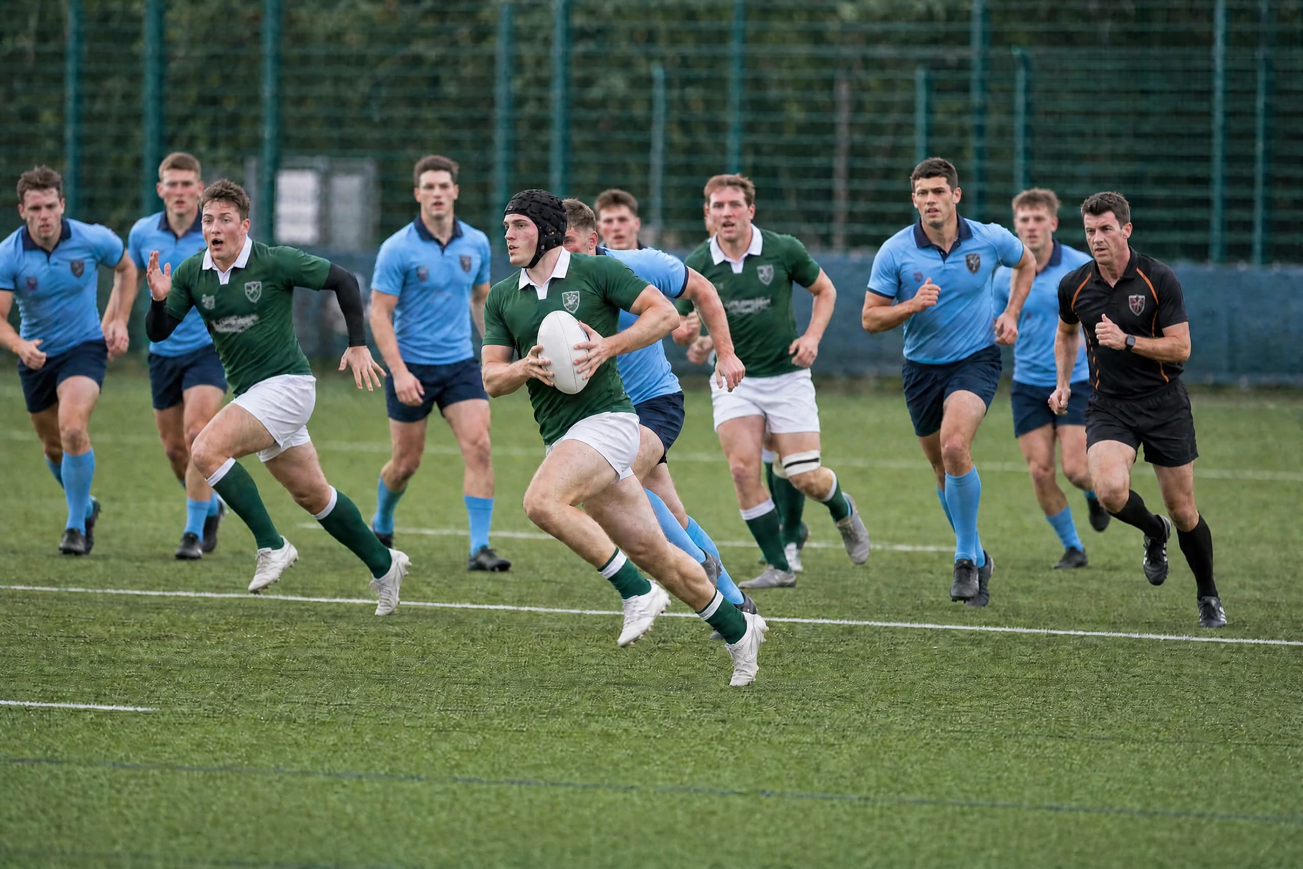 A player in green carrying the ball upfield at pace, with sky-blue defenders and the referee chasing